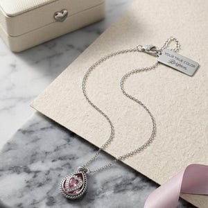 Puede incluir: Un collar plateado con un colgante en forma de l&aacute;grima con una piedra rosa en forma de coraz&oacute;n y piedras m&aacute;s peque&ntilde;as. El collar tiene una cadena delicada y una etiqueta que dice "YOUR TRUE COLOR Brighton". Una peque&ntilde;a caja de joyer&iacute;a crema est&aacute; en el fondo.