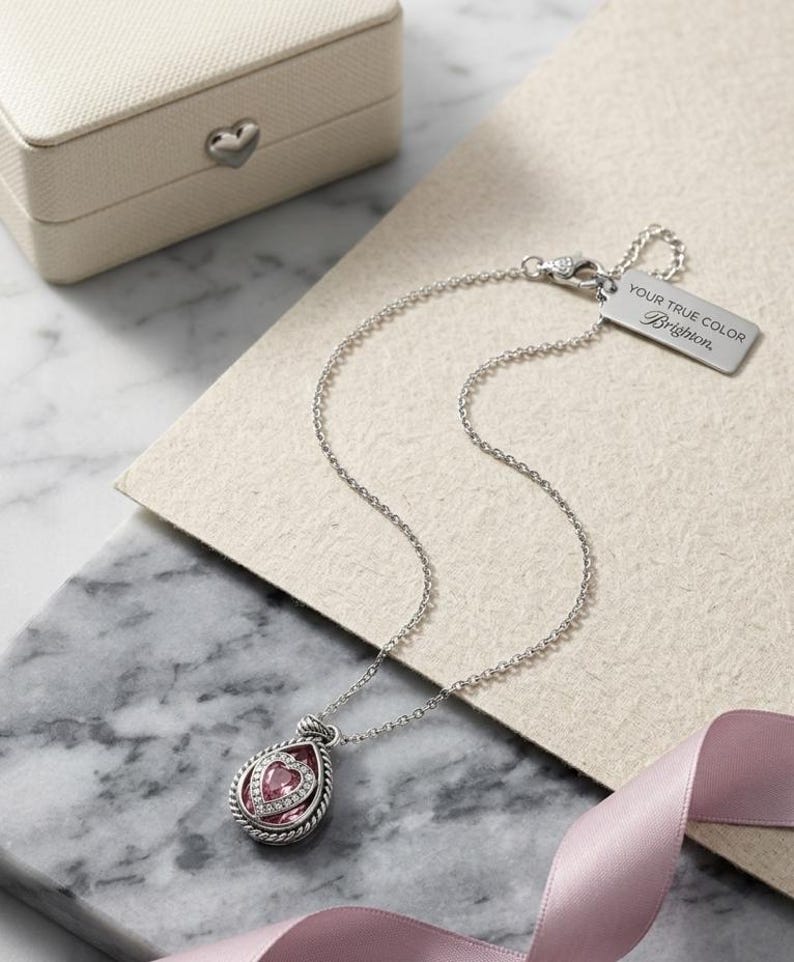 Puede incluir: Un collar plateado con un colgante en forma de l&aacute;grima con una piedra rosa en forma de coraz&oacute;n y piedras m&aacute;s peque&ntilde;as. El collar tiene una cadena delicada y una etiqueta que dice "YOUR TRUE COLOR Brighton". Una peque&ntilde;a caja de joyer&iacute;a crema est&aacute; en el fondo.