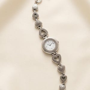 May include: A silver-tone analogue watch featuring a round white face with black hands and markers. The watch band is composed of multiple heart-shaped links with intricate embossed detailing, secured by a toggle clasp at each end of the bracelet.