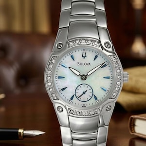 Puede incluir: Un reloj de pulsera Bulova con una correa y caja de acero inoxidable plateado. La esfera es blanca con detalles azules y una pequeña subesfera. El bisel está adornado con pequeñas piedras brillantes. El reloj se muestra sobre una superficie de madera.
