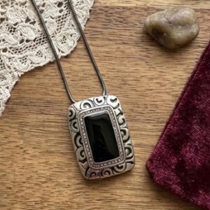 Puede incluir: Un collar con colgante plateado con una piedra negra rectangular. El colgante presenta un dise&ntilde;o ornamental y ondulado en los bordes y cuelga de una fina cadena plateada. El collar est&aacute; sobre una superficie de madera.