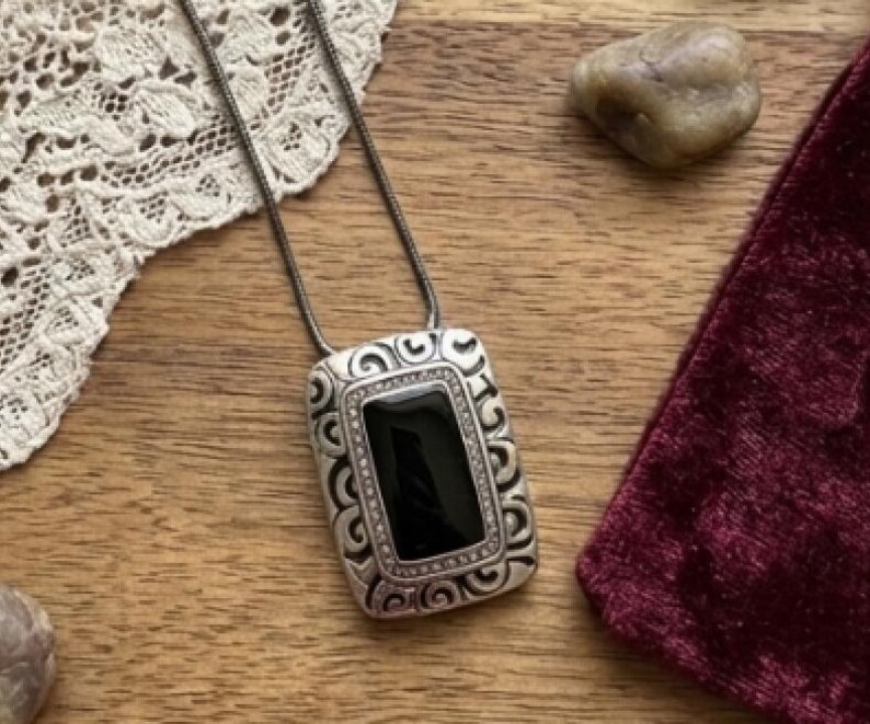 Puede incluir: Un collar con colgante plateado con una piedra negra rectangular. El colgante presenta un dise&ntilde;o ornamental y ondulado en los bordes y cuelga de una fina cadena plateada. El collar est&aacute; sobre una superficie de madera.