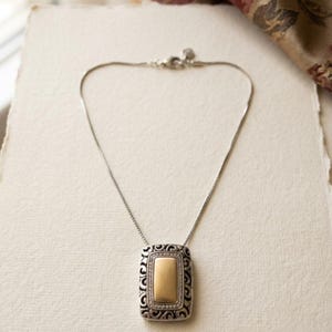 Puede incluir: Collar plateado con un colgante rectangular. El colgante presenta un centro dorado enmarcado por un dise&ntilde;o de filigrana plateada y peque&ntilde;as piedras transparentes. El collar se muestra sobre una superficie blanca.