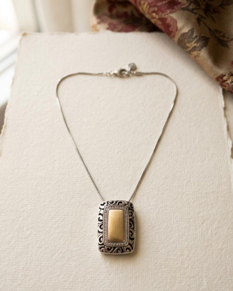Puede incluir: Collar plateado con un colgante rectangular. El colgante presenta un centro dorado enmarcado por un dise&ntilde;o de filigrana plateada y peque&ntilde;as piedras transparentes. El collar se muestra sobre una superficie blanca.