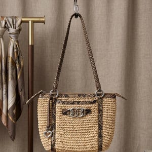 May include: A woven straw handbag featuring brown snakeskin-patterned leather trim, a braided shoulder strap, and a silver-toned metal emblem on the front. A small silver heart-shaped charm hangs from a leather cord attached to the side ring.