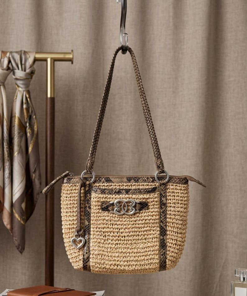 May include: A woven straw handbag featuring brown snakeskin-patterned leather trim, a braided shoulder strap, and a silver-toned metal emblem on the front. A small silver heart-shaped charm hangs from a leather cord attached to the side ring.
