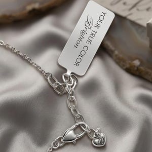 Puede incluir: Un collar plateado con un cierre en forma de coraz&oacute;n y un peque&ntilde;o dije con la letra "B". Una etiqueta rectangular con las palabras "YOUR TRUE COLOR Brighton" est&aacute; adjunta. El collar descansa sobre una superficie de sat&eacute;n gris.