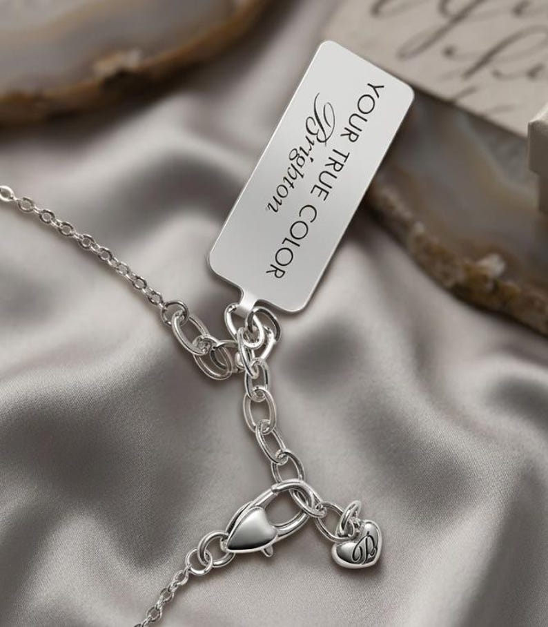 Puede incluir: Un collar plateado con un cierre en forma de coraz&oacute;n y un peque&ntilde;o dije con la letra "B". Una etiqueta rectangular con las palabras "YOUR TRUE COLOR Brighton" est&aacute; adjunta. El collar descansa sobre una superficie de sat&eacute;n gris.