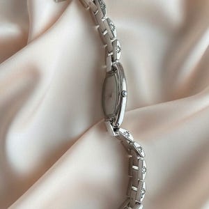 May include: A silver-toned wristwatch featuring an oval-shaped face and a link bracelet. Each individual link is decorated with a small, dark, engraved floral pattern. The watch includes a small crown on the side for time adjustment.