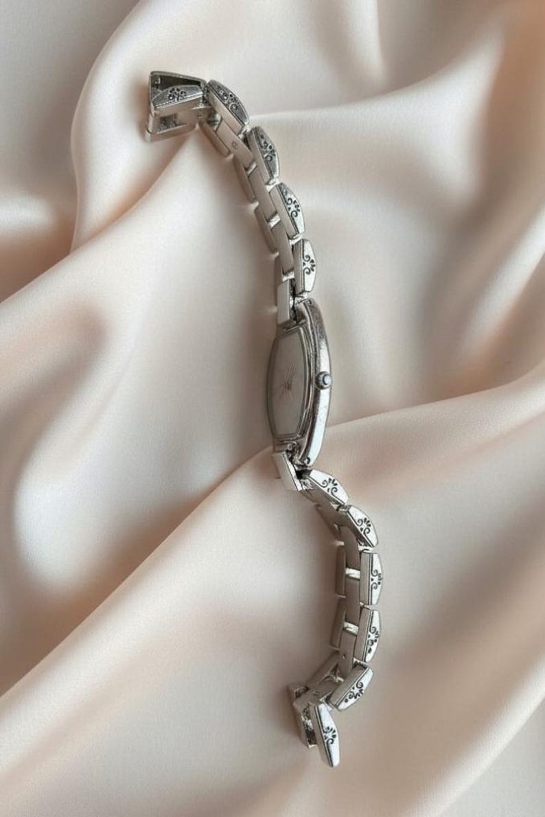 May include: A silver-toned wristwatch featuring an oval-shaped face and a link bracelet. Each individual link is decorated with a small, dark, engraved floral pattern. The watch includes a small crown on the side for time adjustment.