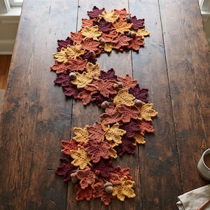 Può includere: Un runner da tavola decorativo composto da numerose foglie d'acero all'uncinetto in tonalità bordeaux intenso, arancione bruciato, giallo senape e marrone. Le foglie sono disposte in un motivo organico sinuoso e intervallate da piccole ghiande.