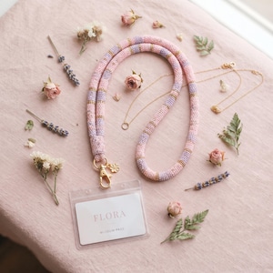 May include: A beaded lanyard featuring a repeating pattern of soft pink, lavender, and metallic gold beads. The lanyard includes a gold-toned metal lobster clasp attached to a clear plastic badge holder containing a card labelled FLORA MUSEUM PASS.