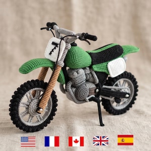 Könnte beinhalten: Ein handgefertigtes Häkel-Motorrad-Spielzeug mit grünem Tank und Schutzblechen, schwarzem Sitz und einem weißen vorderen Nummernschild mit der schwarzen Ziffer 7. Das Modell hat beige Vordergabeln, graue Motordetails und schwarze Stollenreifen.