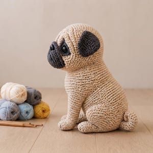 Könnte beinhalten: Ein gehäkeltes Mops-Spielzeug aus beigem Garn mit dunkelgrauer Schnauze, Ohren und großen schwarzen Kunststoffaugen. Das Stofftier sitzt in einer Profilansicht mit einem kleinen geringelten Schwanz, begleitet von mehreren Garnknäueln und Häkelnadeln.
