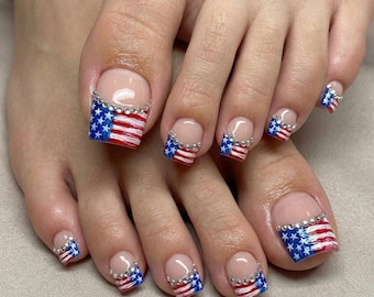 Puede incluir: Uñas de los pies con estilo de manicura francesa y un diseño patriótico de la bandera estadounidense en las puntas. Las puntas están pintadas con rayas rojas y blancas y secciones azules con estrellas blancas, acentuadas por una fila de pedrería plateada.