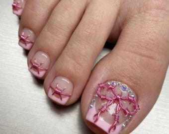 Puede incluir: Primer plano de un pie con un diseño de uñas rosa y transparente. Cada uña del pie está pintada con una punta rosa y presenta un diseño de alambre de púas rosa. El dedo gordo tiene un borde decorativo de pedrería.