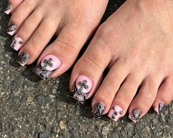 Puede incluir: Primer plano de pies con esmalte de uñas rosa y arte de uñas intrincado. Los dedos gordos presentan un diseño de cruz negra sobre un patrón de encaje. Otras uñas tienen encaje, tachuelas y diseños de cruz.
