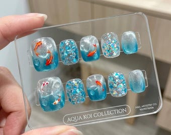 Puede incluir: Un juego de diez uñas postizas de la colección Aqua Koi con un degradado azul y transparente, ilustraciones de peces koi naranjas y blancos, purpurina plateada y copos de lámina iridiscente azul sobre una tarjeta expositora transparente.