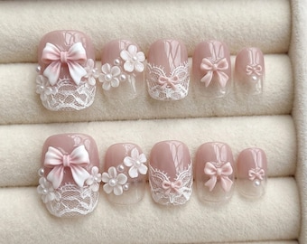 Puede incluir: Un conjunto de diez uñas artificiales en un tono rosa claro. Cada uña presenta elementos decorativos como lazos, encaje y detalles florales. Las uñas están dispuestas en dos filas de cinco, mostrando los diferentes diseños.