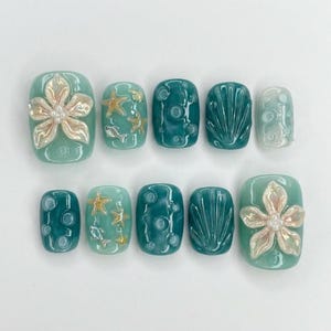 Könnte beinhalten: Ein Set aus zehn künstlichen Nägeln in verschiedenen Türkis- und Seegrün-Tönen. Einige Nägel weisen 3D-Verzierungen mit Blumen, Seesternen und Muscheln auf. Andere Nägel haben ein strukturiertes Design mit kleinen Kreisen oder Muschelmustern.