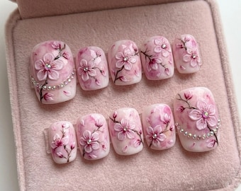 Puede incluir: Un conjunto de diez uñas artificiales con un diseño floral rosa. Cada uña presenta un diseño de flor de cerezo en 3D con un acento de pedrería. Las uñas se muestran en una caja de terciopelo rosa.