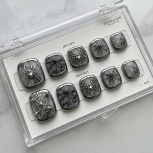 Könnte beinhalten: Ein Set aus künstlichen Nägeln mit Spinnen- und Spinnennetz-Design. Die Nägel sind grau und silberfarben, mit glänzendem Finish. Das Set befindet sich in einer transparenten Kunststoffbox mit dem Text "SPIDER & WEB COLLECTIVE".