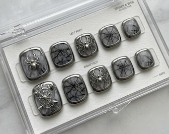 Puede incluir: Un juego de uñas postizas con un diseño de araña y telaraña. Las uñas son grises y plateadas, con un acabado brillante. El juego está en una caja de plástico transparente, con el texto "SPIDER & WEB COLLECTIVE".
