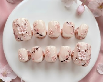 Puede incluir: Un juego de diez uñas postizas con una base rosa pálido brillante, ramas de cerezo pintadas a mano en marrón oscuro, pétalos de flores rosas esculpidos en tres dimensiones y pequeñas perlas blancas dispuestas en un patrón floral.