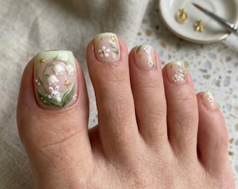 Puede incluir: Uñas de los pies con una base de color verde claro y nude, decoradas con flores de lirio de los valles pintadas a mano, hojas verdes y un pequeño conejo blanco en el dedo gordo. El diseño incluye diminutas estrellas doradas y flores blancas.