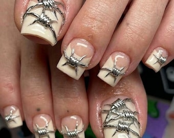 Puede incluir: Primer plano de un pie con manicura con uñas cuadradas. Cada uña está pintada con un color beige claro y adornada con diseños de alambre de púas plateado. Las uñas tienen un diseño de punta francesa.
