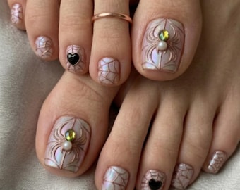 Puede incluir: Primer plano de pies con pedicura y arte de uñas intrincado. Las uñas de los pies presentan un diseño de telaraña con acentos de araña y gemas. Un anillo de oro rosa adorna un dedo del pie. Las uñas están pintadas en un color nacarado.