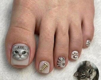 Puede incluir: Uñas de los pies decoradas con temática felina. El dedo gordo presenta una cara de gato atigrado gris en estilo 3D. Otros dedos muestran una espina de pescado beige, patrones de pelaje, una huella marrón y rayas. Texto: TABBY CAT INSPIRATION.