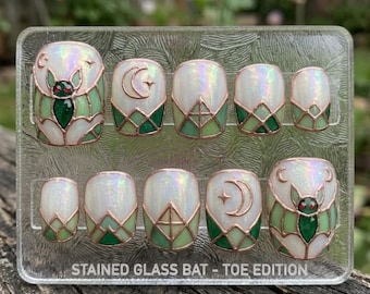 Puede incluir: Un juego de uñas postizas para los dedos de los pies con un diseño de murciélago de vidriera. Las uñas presentan una base blanca nacarada con detalles verdes y cobrizos. El texto "STAINED GLASS BAT - TOE EDITION" está impreso en el embalaje de plástico transparente.