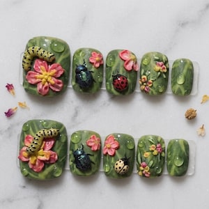 May include: A set of ten press-on nails featuring a green marbled base with three-dimensional embellishments including yellow caterpillars, pink flowers, iridescent blue beetles, red and yellow ladybugs, and clear dew drop accents on each nail.