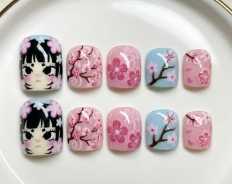 Puede incluir: Un juego de diez uñas postizas con acabado brillante. Dos uñas muestran una ilustración estilizada de un personaje con cabello negro y flores rosas. Las uñas restantes presentan varios diseños de flores de cerezo rosas sobre fondos azul claro y rosa.