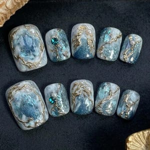 May include: A set of ten artificial nails with a marbled blue and white design. Each nail features gold-colored, swirling accents, and some are adorned with pearls or teal-colored gems. The nails have a glossy finish.