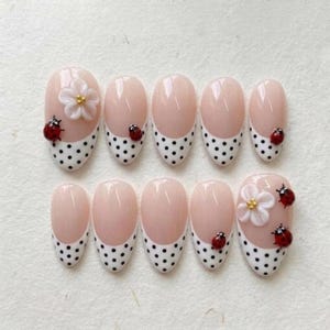 May include: A set of ten press-on nails featuring a French manicure style with a nude base and white tips adorned with black polka dots. Several nails are decorated with small, three-dimensional white flowers and tiny red ladybug accents.