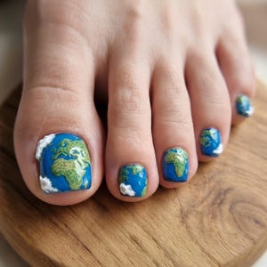 May include: Toenails painted with a detailed globe design featuring blue oceans, green landmasses, and white clouds. The artwork covers each nail, depicting continents and swirling cloud formations in a glossy finish across all five visible toes.