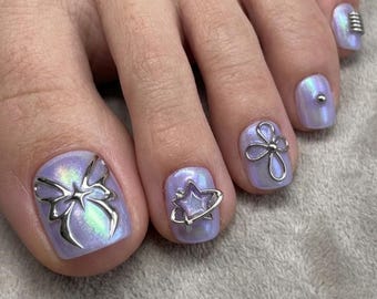 Puede incluir: Uñas de los pies pintadas con esmalte violeta claro brillante, con dijes de metal plateado. El dedo gordo tiene un dije de lazo, el segundo una estrella, el tercero una flor, el cuarto un pequeño remache y el dedo meñique un anillo.