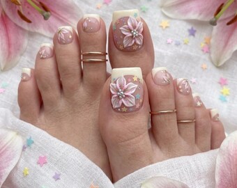 Puede incluir: Un conjunto de uñas de los pies con base color crema y detalles florales rosas en relieve en los dedos gordos, decoradas con pequeñas cuentas plateadas y estrellas de colores pastel dispersas. El diseño incluye finos anillos dorados.