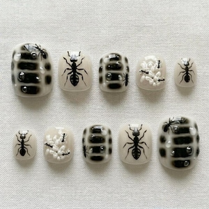 May include: A set of ten artificial nails with a unique ant-themed design. The nails feature a combination of black and white patterns, with some adorned with detailed ant illustrations and small, decorative embellishments. The nails are in various sizes.