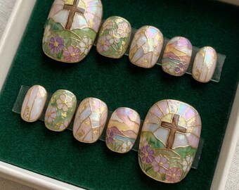 Puede incluir: Un conjunto de uñas postizas con temática religiosa. Las uñas presentan un diseño de cruz, detalles florales y un efecto de vidriera con colores pastel. Las uñas están dispuestas sobre una lámina de plástico transparente.
