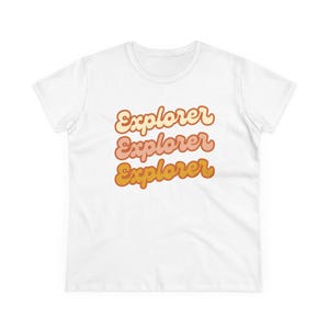 May include: White t-shirt with the word "Explorer" in a retro font, repeated three times in shades of peach, pink, and gold. The shirt is a crew neck and appears to be made of cotton.