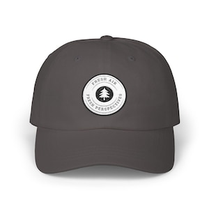 May include: A charcoal gray baseball cap with a circular patch featuring a pine tree graphic. The patch has the words "FRESH AIR" above the tree and "FRESH PERSPECTIVES" below it, all in a circular design.