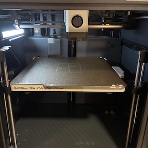May include: A 3D printer with a black and gray interior. The build plate is a textured gray, with a warning label and the dimensions 256x256x256mm. The printer's components include a print head, support rods, and a control panel.