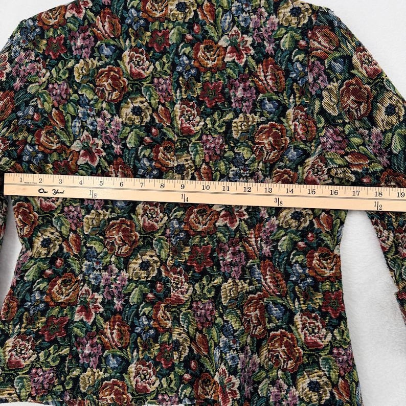 May include: A floral tapestry jacket with a colorful pattern of red, green, and purple flowers. The jacket has long sleeves and a tailored fit. A wooden ruler is placed across the jacket, measuring approximately 19 inches.
