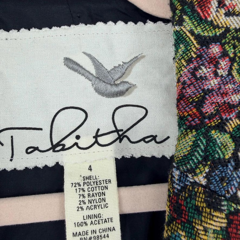 May include: Close-up of a garment label with the brand name "Tabitha" in cursive. The label includes a stitched bird design and fabric composition details: 72% polyester, 17% cotton, 7% rayon, 2% nylon, and 2% acrylic. The lining is 100% acetate.
