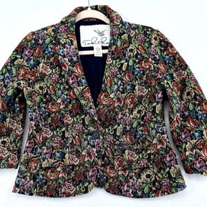 May include: A floral tapestry blazer with a dark navy blue interior. The jacket features a single-button closure and two front pockets. The floral pattern includes red, green, purple, and blue flowers on a black background. The label reads "Tabitha".
