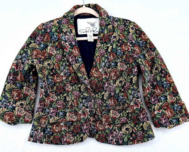 May include: A floral tapestry blazer with a dark navy blue interior. The jacket features a single-button closure and two front pockets. The floral pattern includes red, green, purple, and blue flowers on a black background. The label reads "Tabitha".