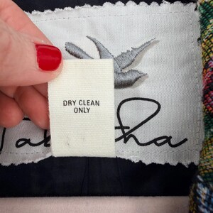 May include: A close-up of a clothing label with the text "DRY CLEAN ONLY". The label is white with a gray bird design and the name "Jatara" in black script. The label is held by a hand with red nail polish.